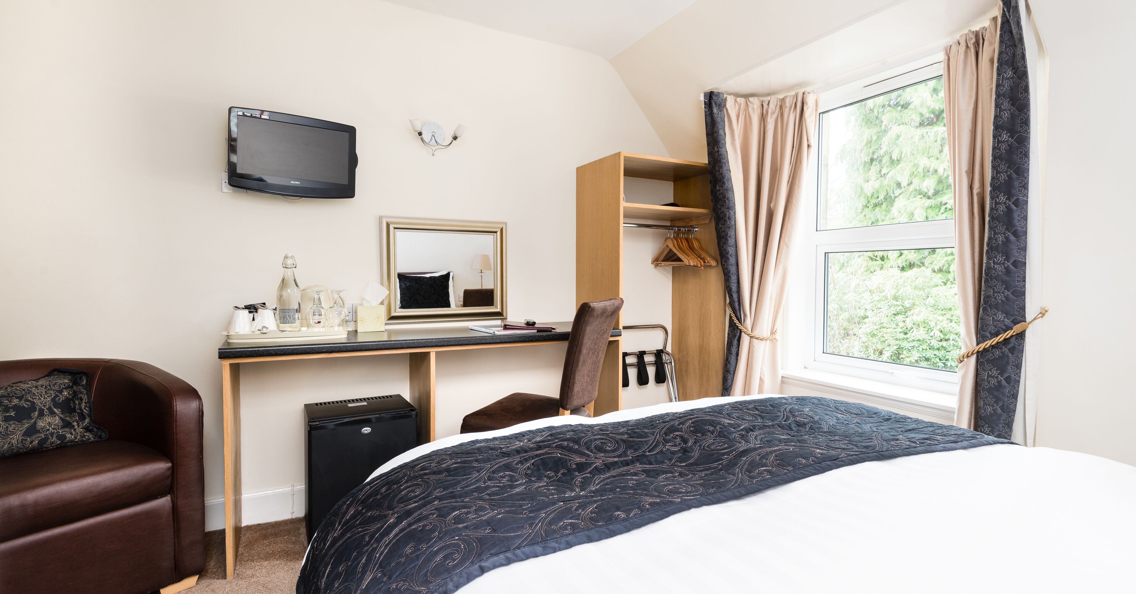 A hotel room with a bed, a desk with a computer, a chair, a TV, a mirror, a window with curtains, and a coat rack.