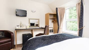 A hotel room with a bed, a desk with a computer, a chair, a TV, a mirror, a window with curtains, and a coat rack.