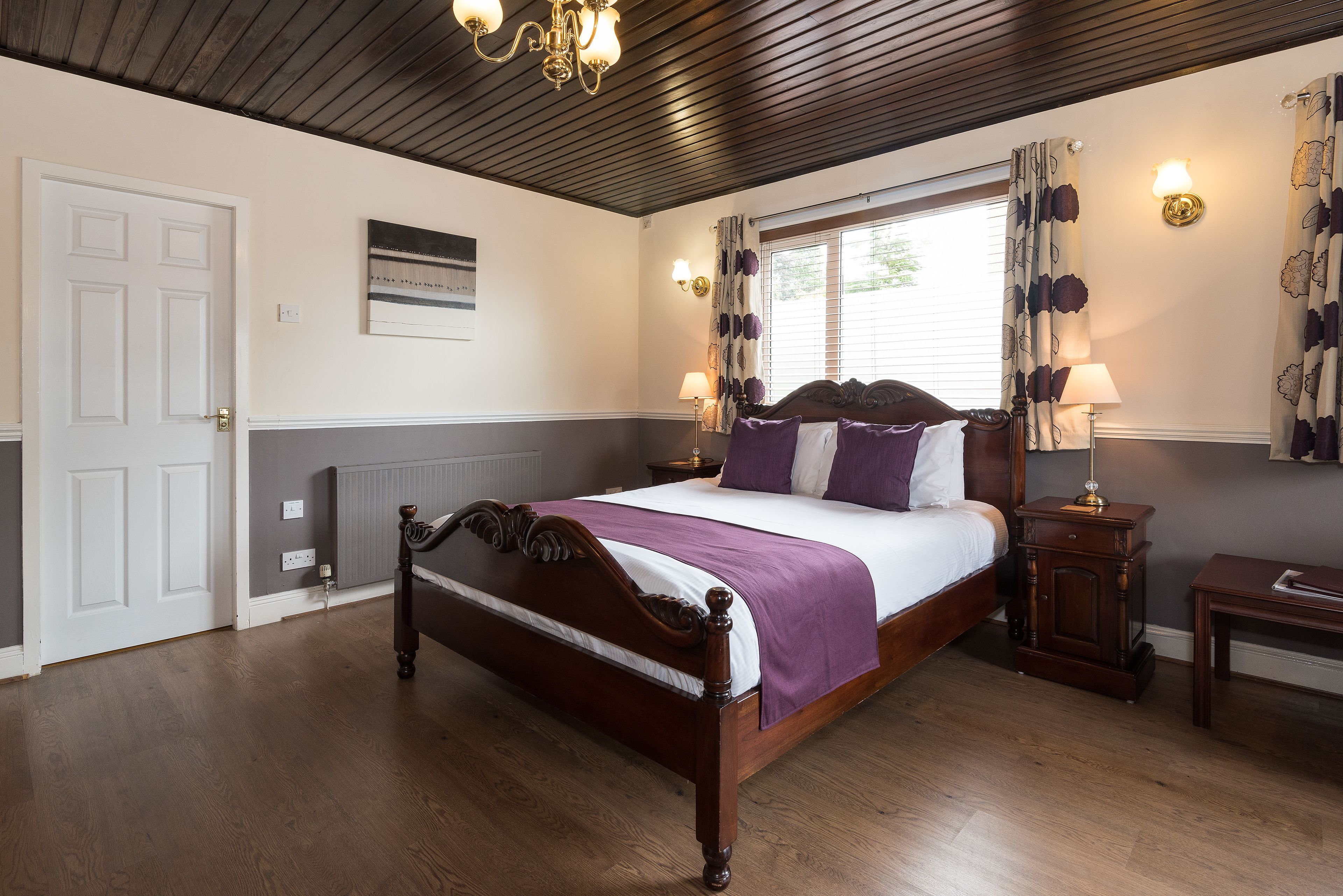 A bedroom with a wooden bed, a nightstand, a lamp, a window with curtains, and a wall-mounted light fixture.