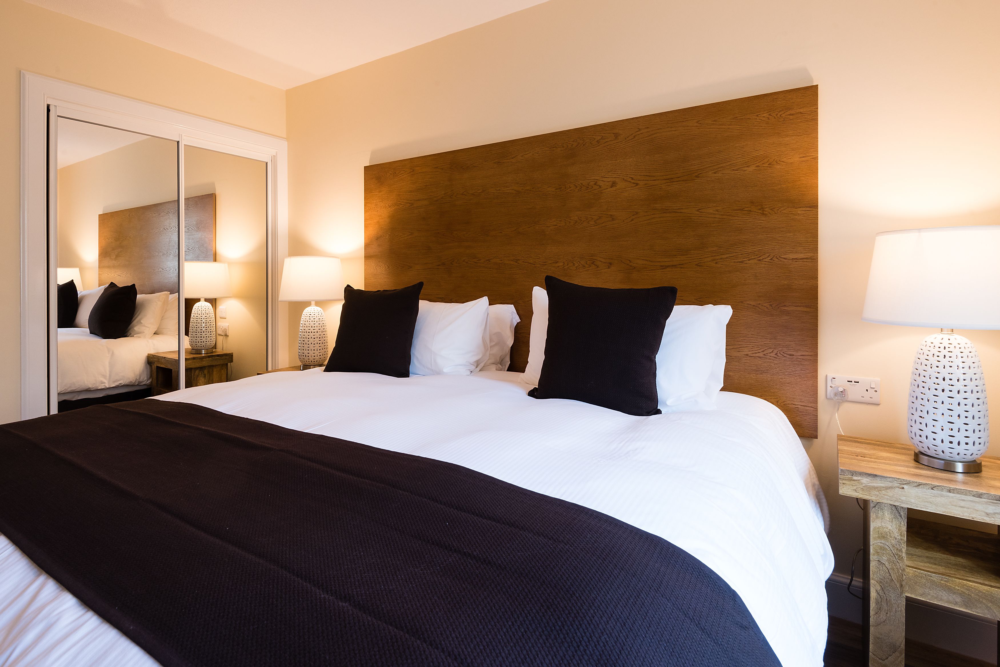 Habitación de hotel con una cama grande, cabecera de madera, dos mesitas de noche con lámparas y un espejo en la pared.