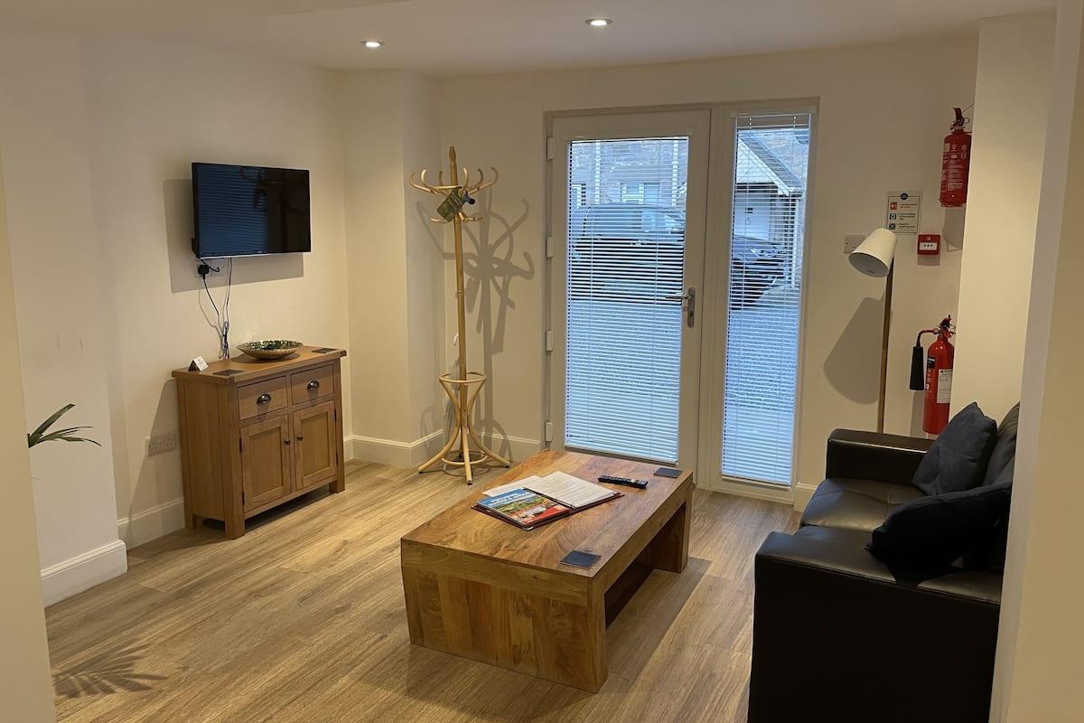 Una sala de estar moderna con una mesa de centro de madera, un televisor de pantalla plana montado en la pared, un mueble de madera y una lámpara de pie.