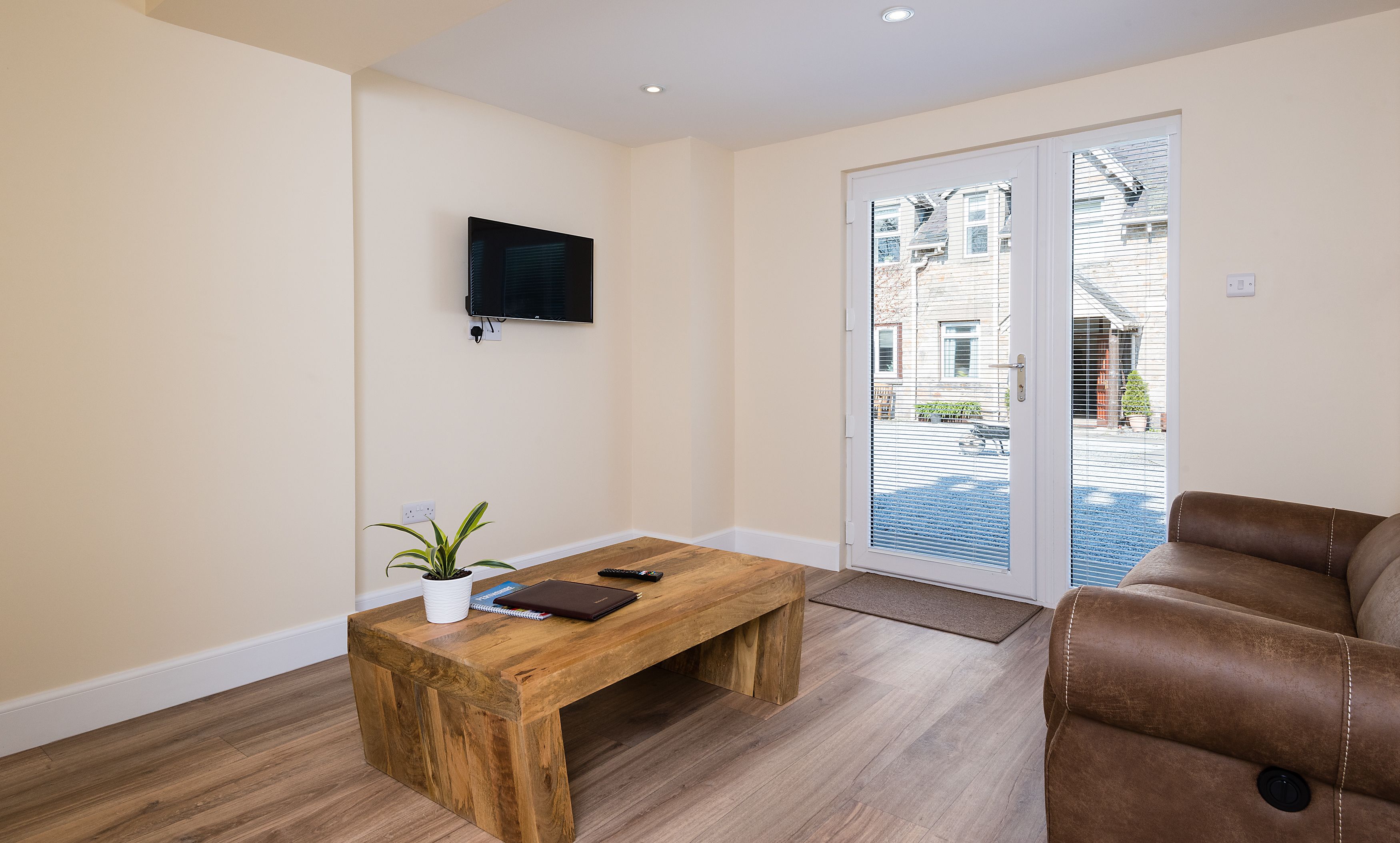 A modern living room with a wooden coffee table, a brown leather sofa, a flat-screen TV mounted on the wall, and a sliding glass door leading to an outdoor area.