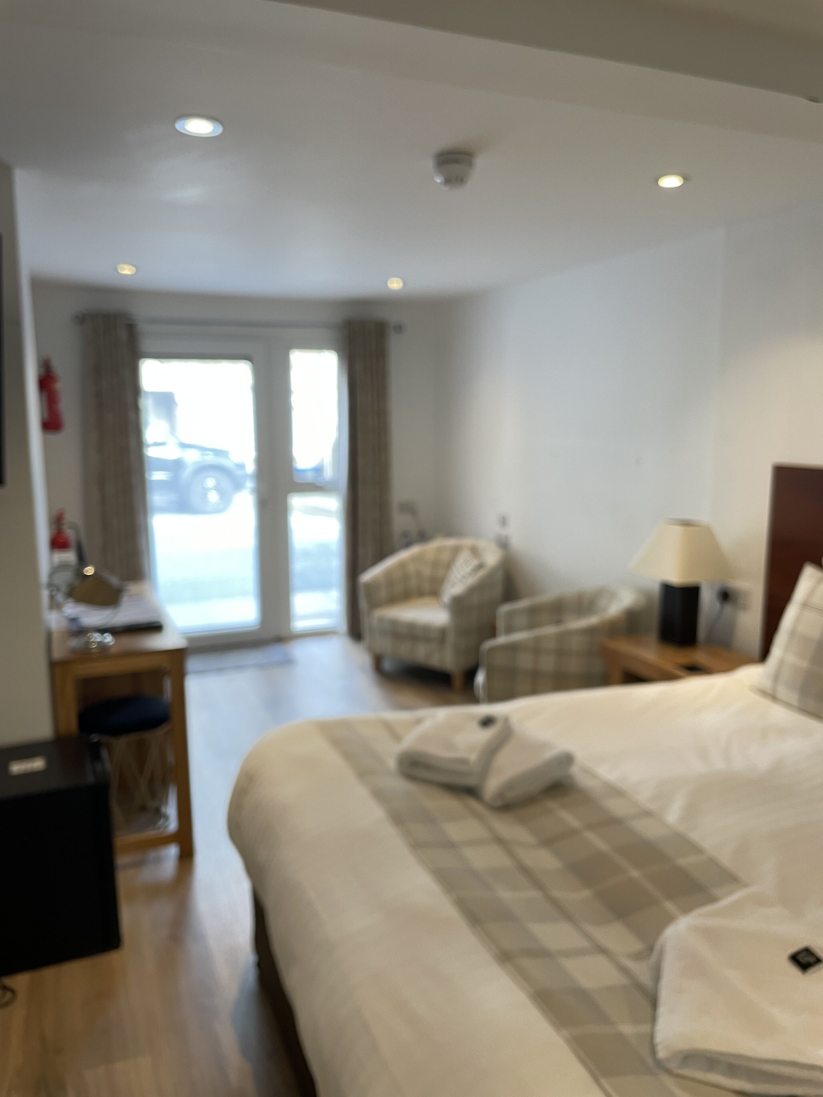 A hotel room with a large bed, two armchairs, a desk with a lamp, and a view of the outside through a glass door.
