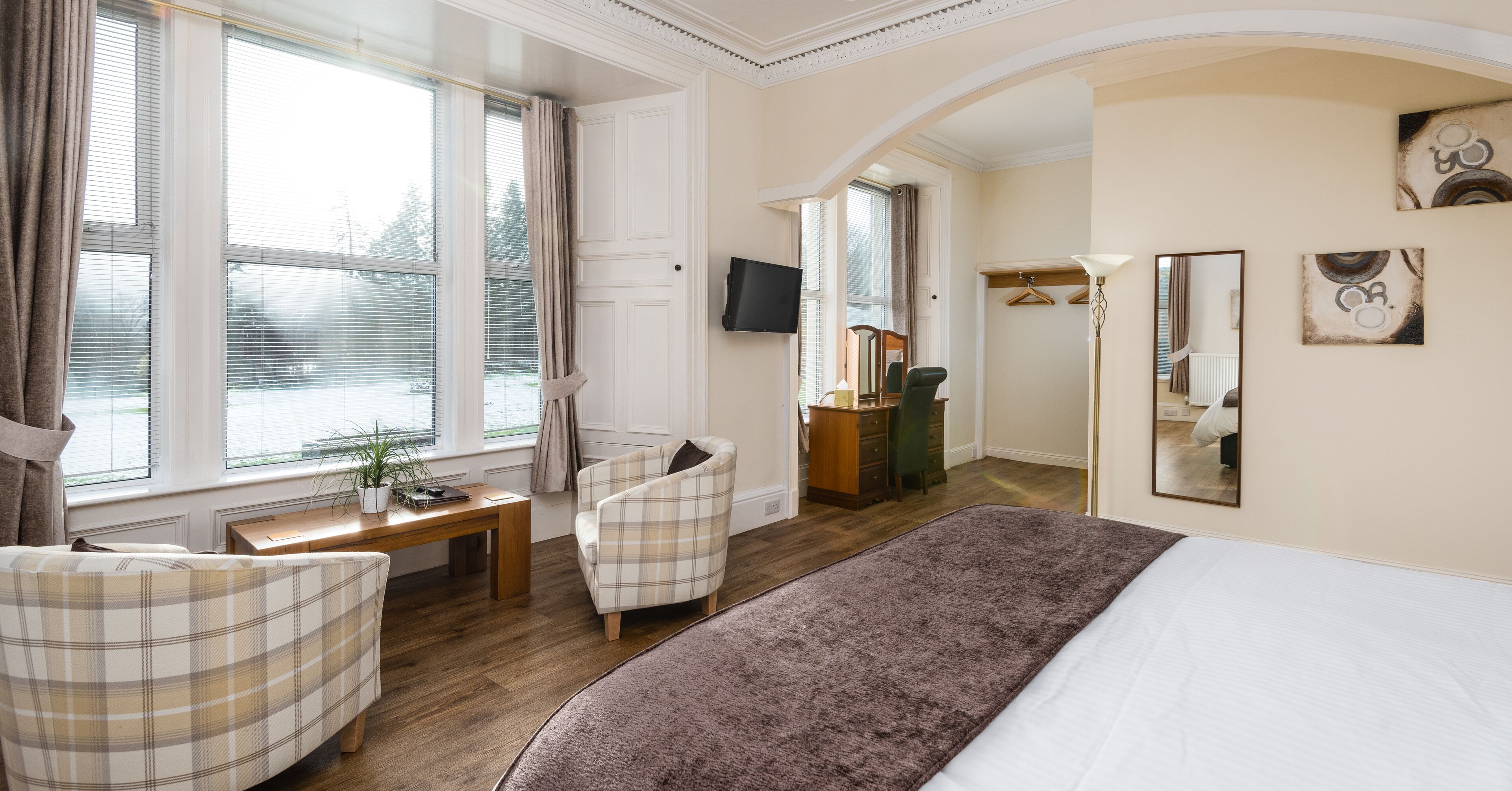 Habitación de hotel con cama, dos sillas, escritorio, televisor y una ventana grande con cortinas.