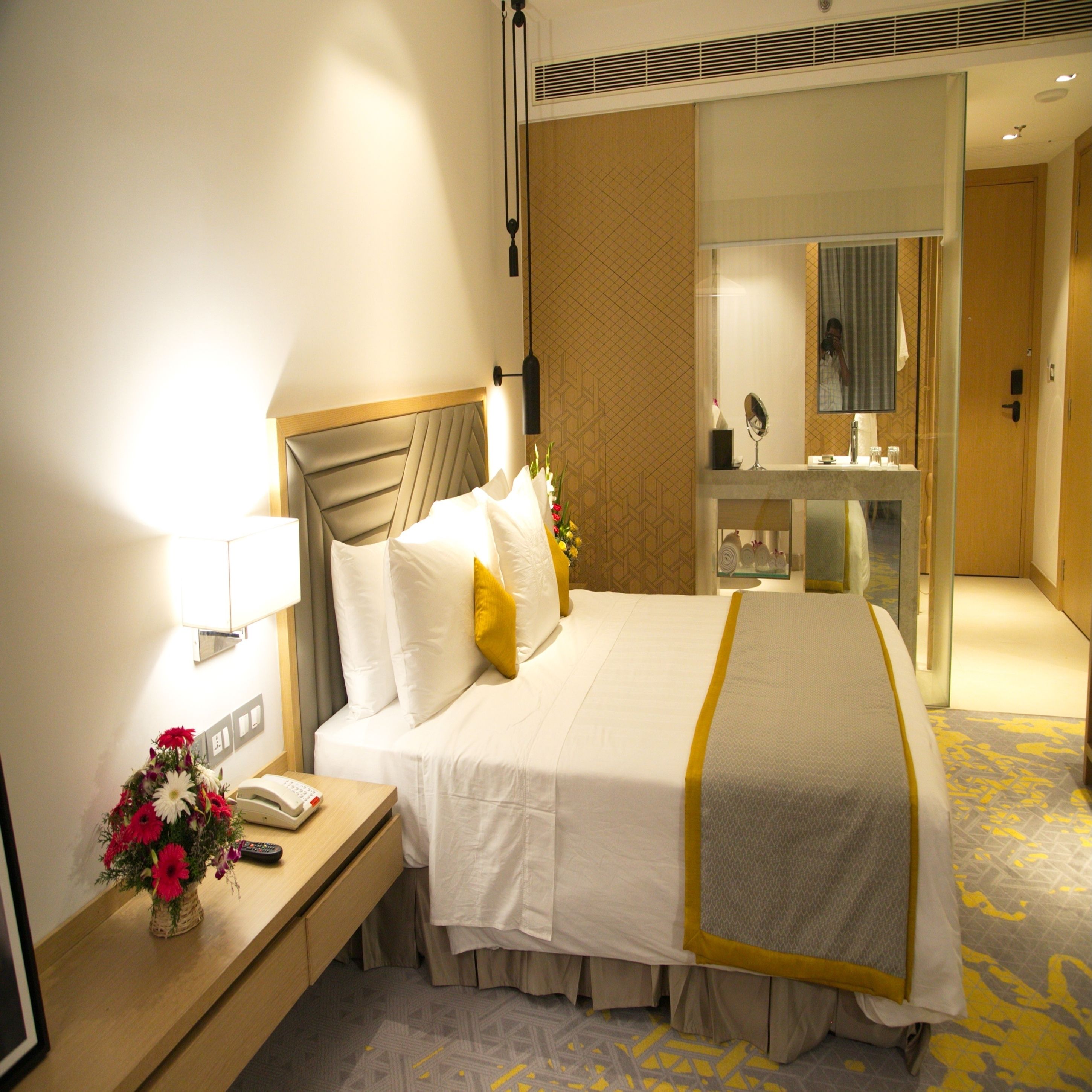Habitación de hotel con una cama bien hecha, una mesita de noche con lámpara, teléfono y un florero con flores.