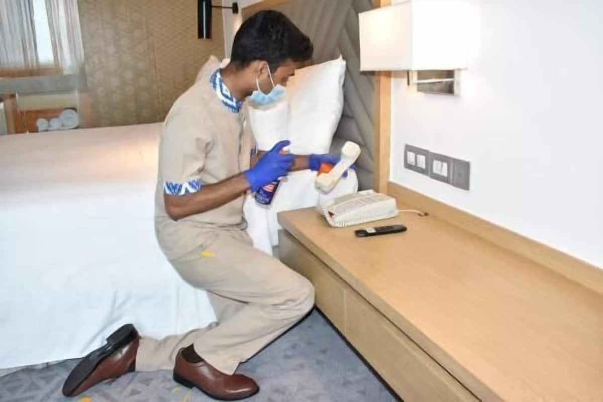 Iemand in een hotelkamer, knielend op de vloer, met een telefoon en fles in de hand, met een bed en nachtkastje op de achtergrond.