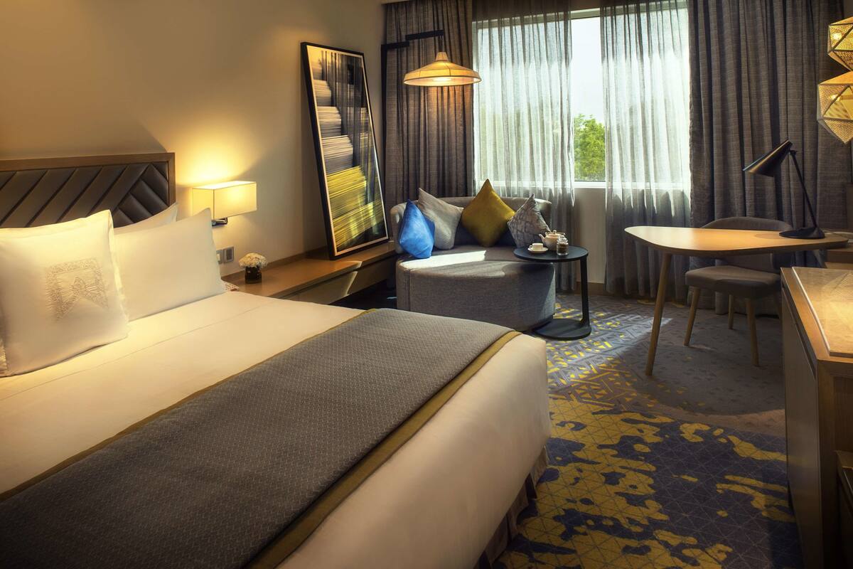 Een hotelkamer met een groot bed, een bureau, een stoel en een bank. Er is een raam met gordijnen en een lamp op het bureau.