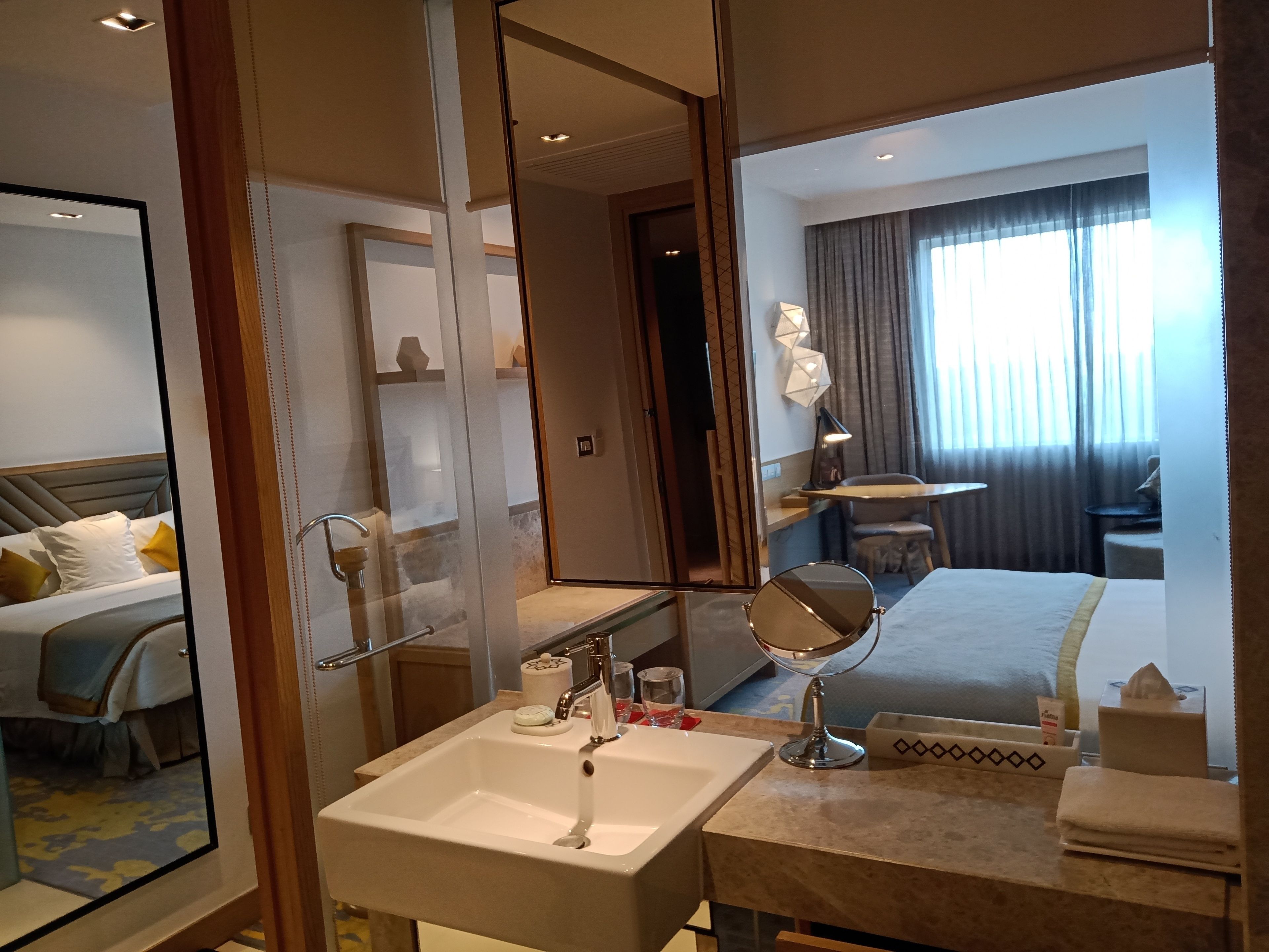 A hotel room with a large mirror, a sink, a bed, a desk, and a window with curtains.