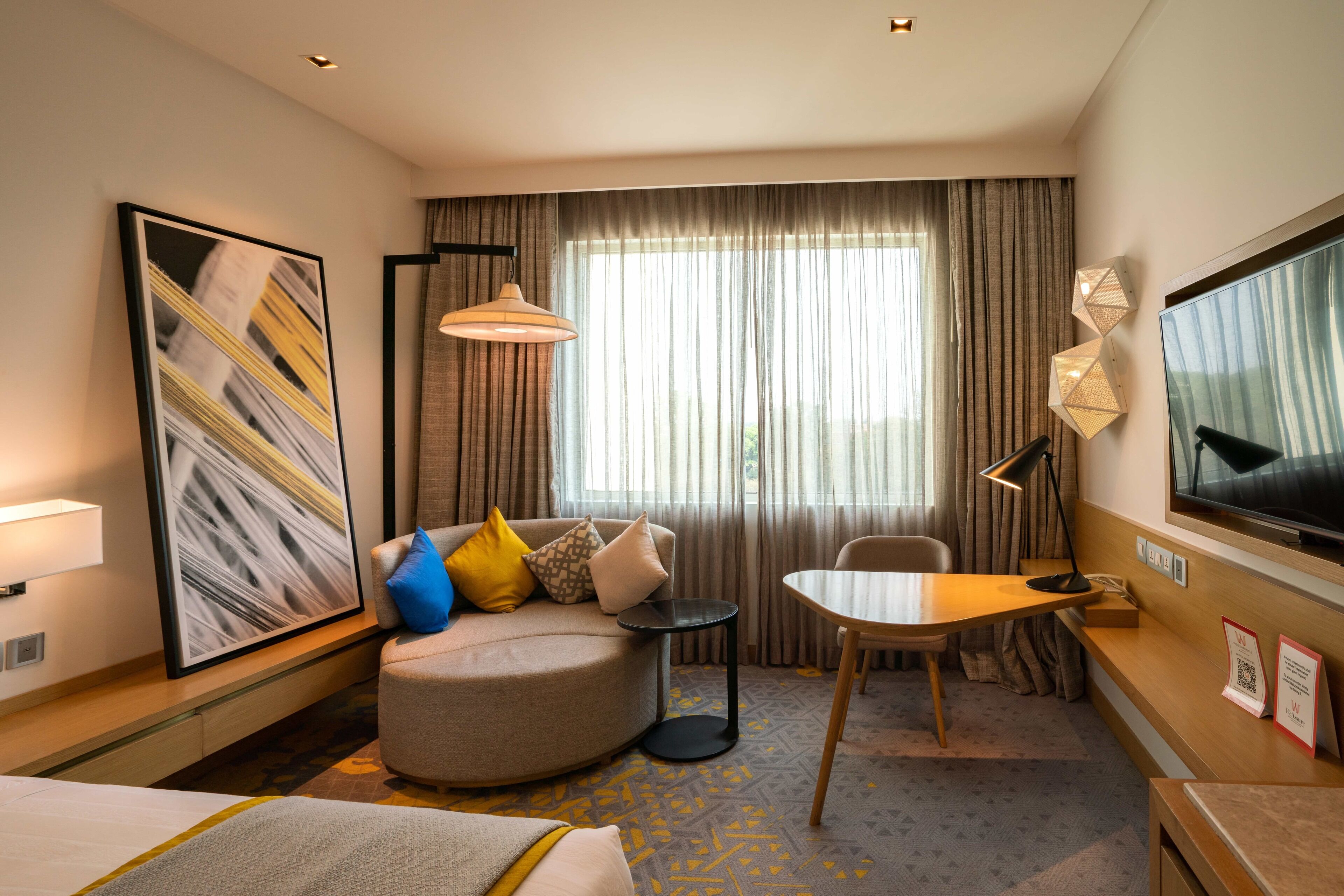 A hotel room with a bed, a round seating area with cushions, a small table, a TV, and a large window with curtains.