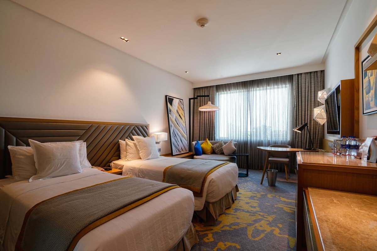 Een hotelkamer met twee bedden, een bureau, een stoel, een lamp en een groot raam met gordijnen.