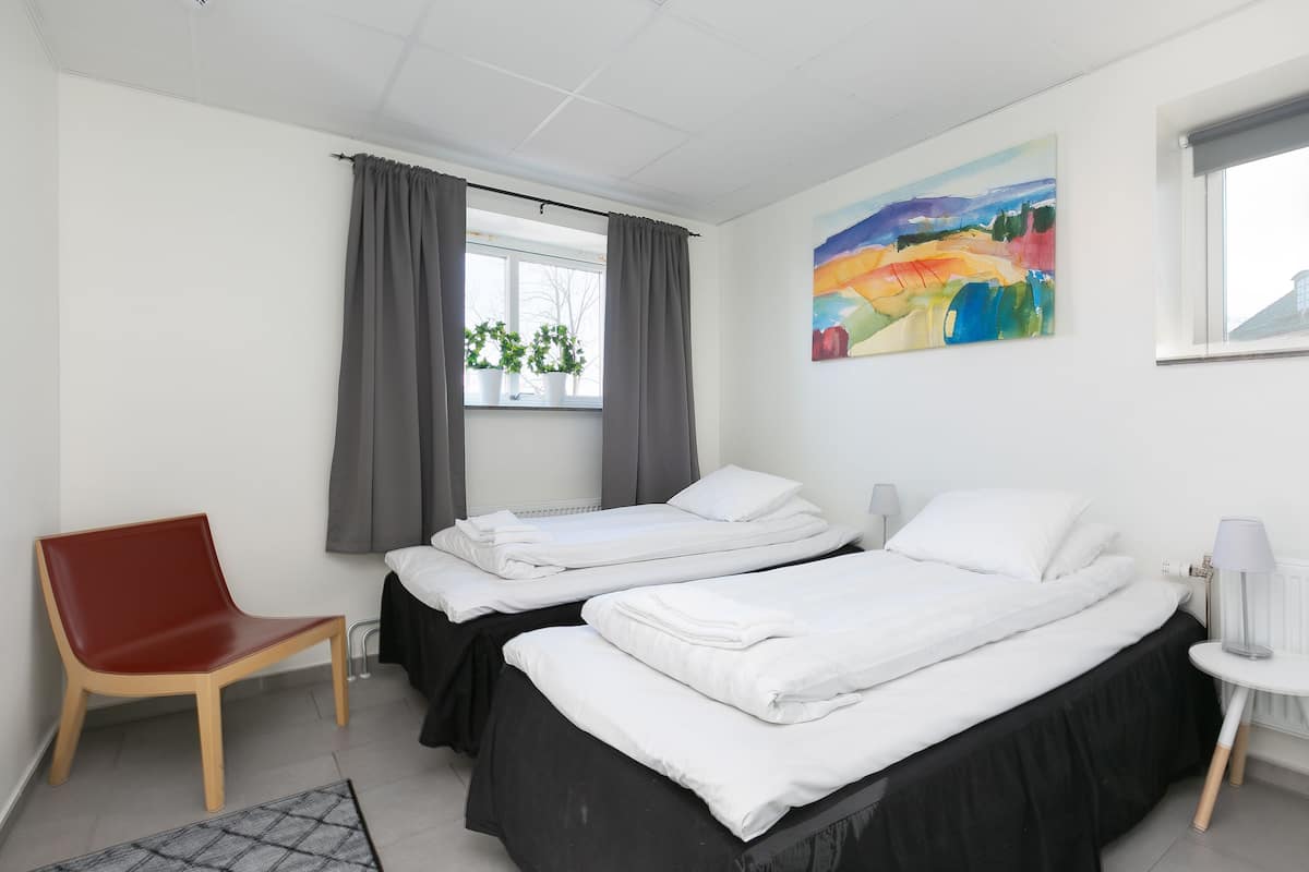 Habitación de hotel con dos camas, una silla roja, una ventana con cortinas, un cuadro en la pared y una mesita con lámpara.