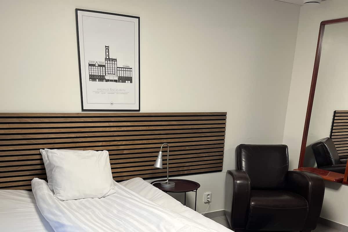 Una cama bien tendida con una toalla doblada, una lámpara de noche, una silla y un cuadro en la pared.