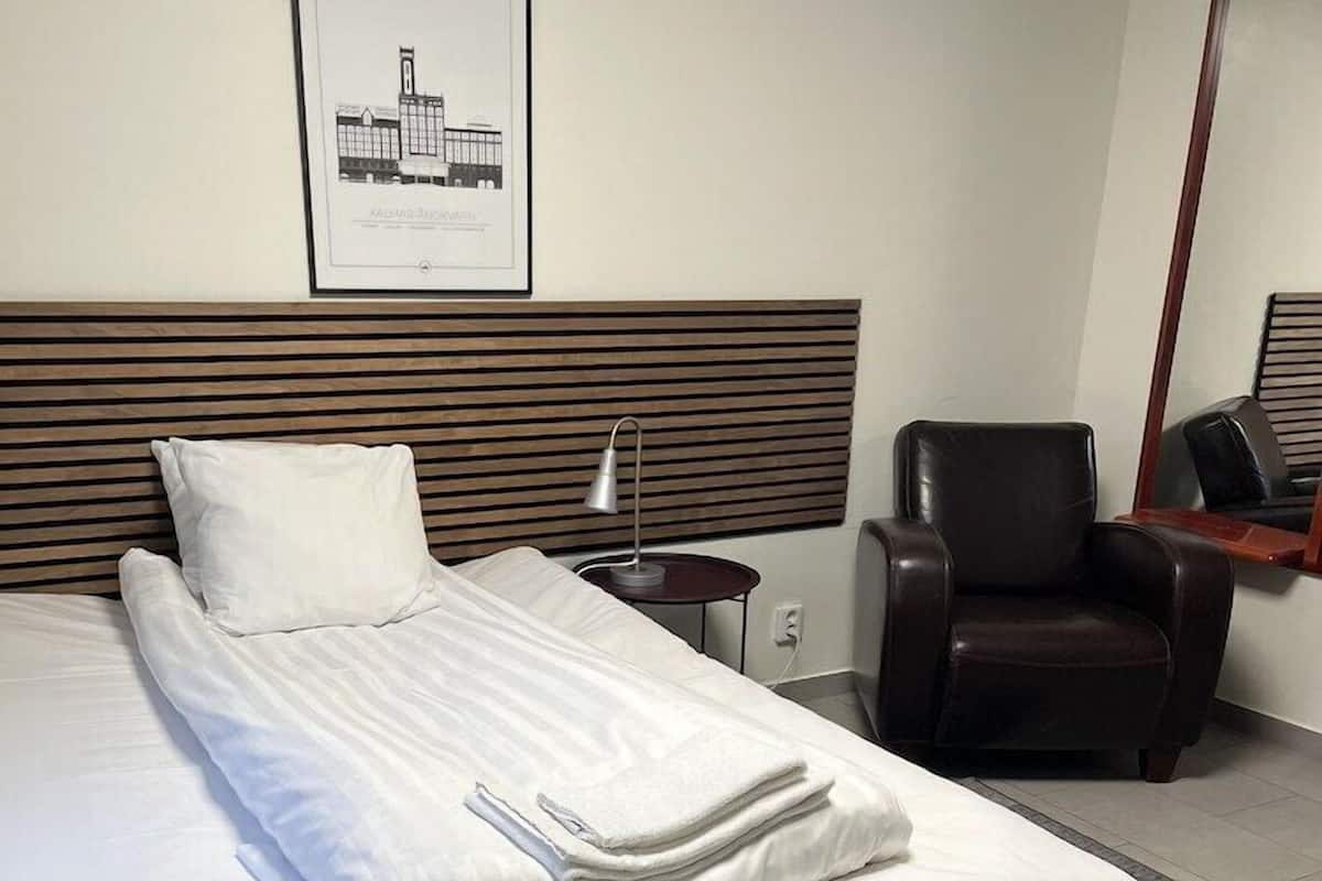 Habitación de hotel con cama bien hecha, una silla, una mesita con lámpara y un cuadro en la pared.
