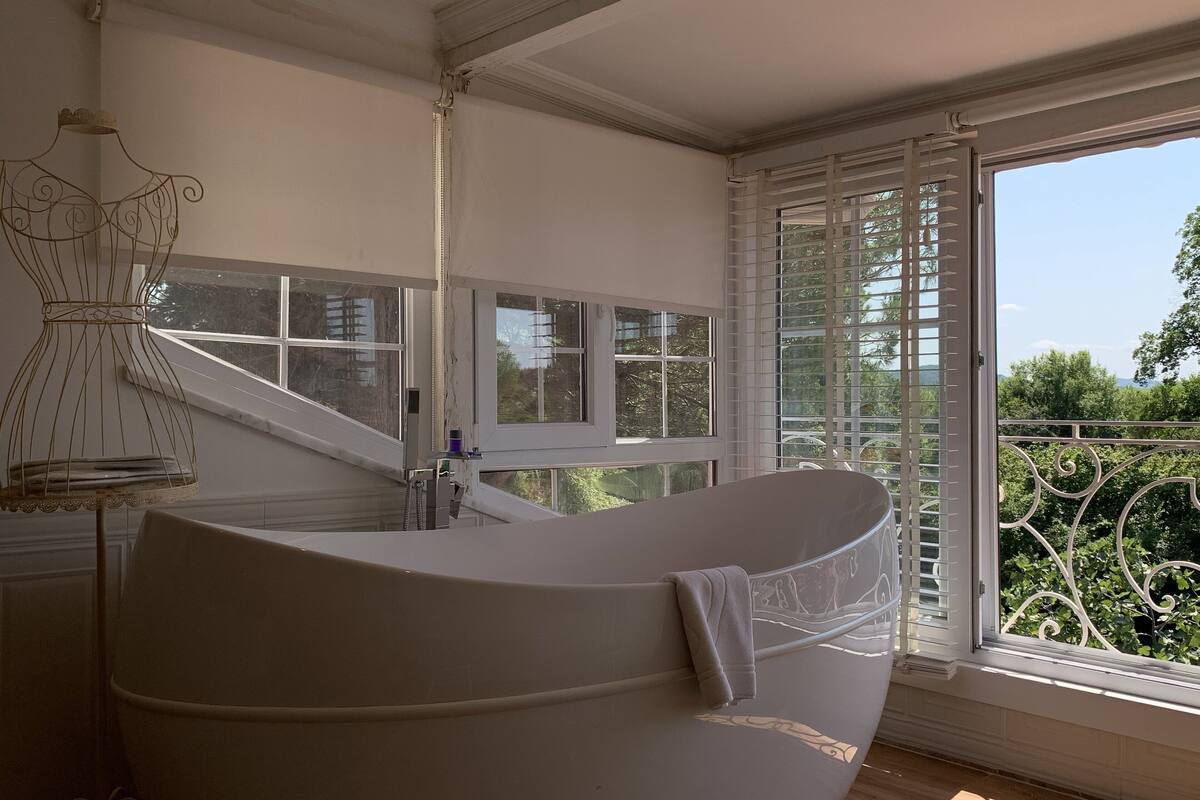 Une pièce avec une grande baignoire, une fenêtre avec des stores et un élément décoratif en métal accroché au mur.