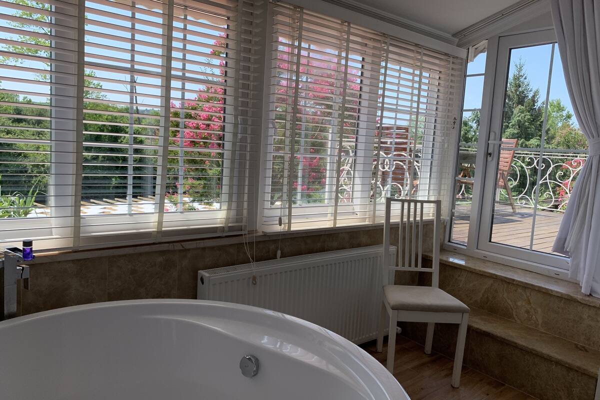 Une salle de bain avec une baignoire, une chaise et une vue sur un jardin grâce à de grandes fenêtres.