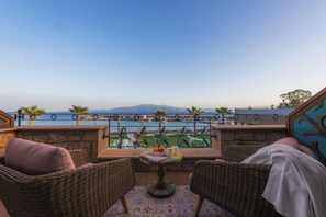 A balcony with wicker chairs, a small table with a fruit bowl, and a view of the sea and mountains. - Assos Zeytinhan Hotel Special Class (Ayvacik)