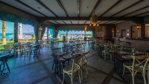 A spacious outdoor dining area with multiple tables and chairs, a view of the sea, and a rustic interior with exposed beams and a brick wall.