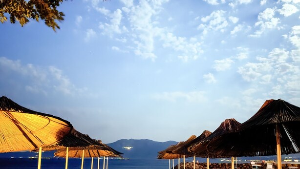 A beachside resort with thatched umbrellas, wooden deck chairs, and a view of the sea and mountains.