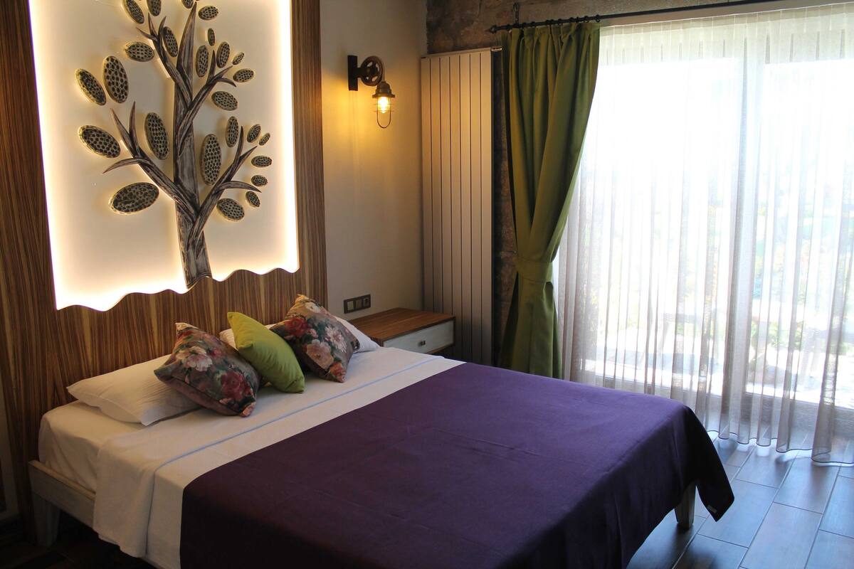 Une chambre à coucher avec un lit, des oreillers, un luminaire fixé au mur, une table de chevet et une fenêtre avec des rideaux.