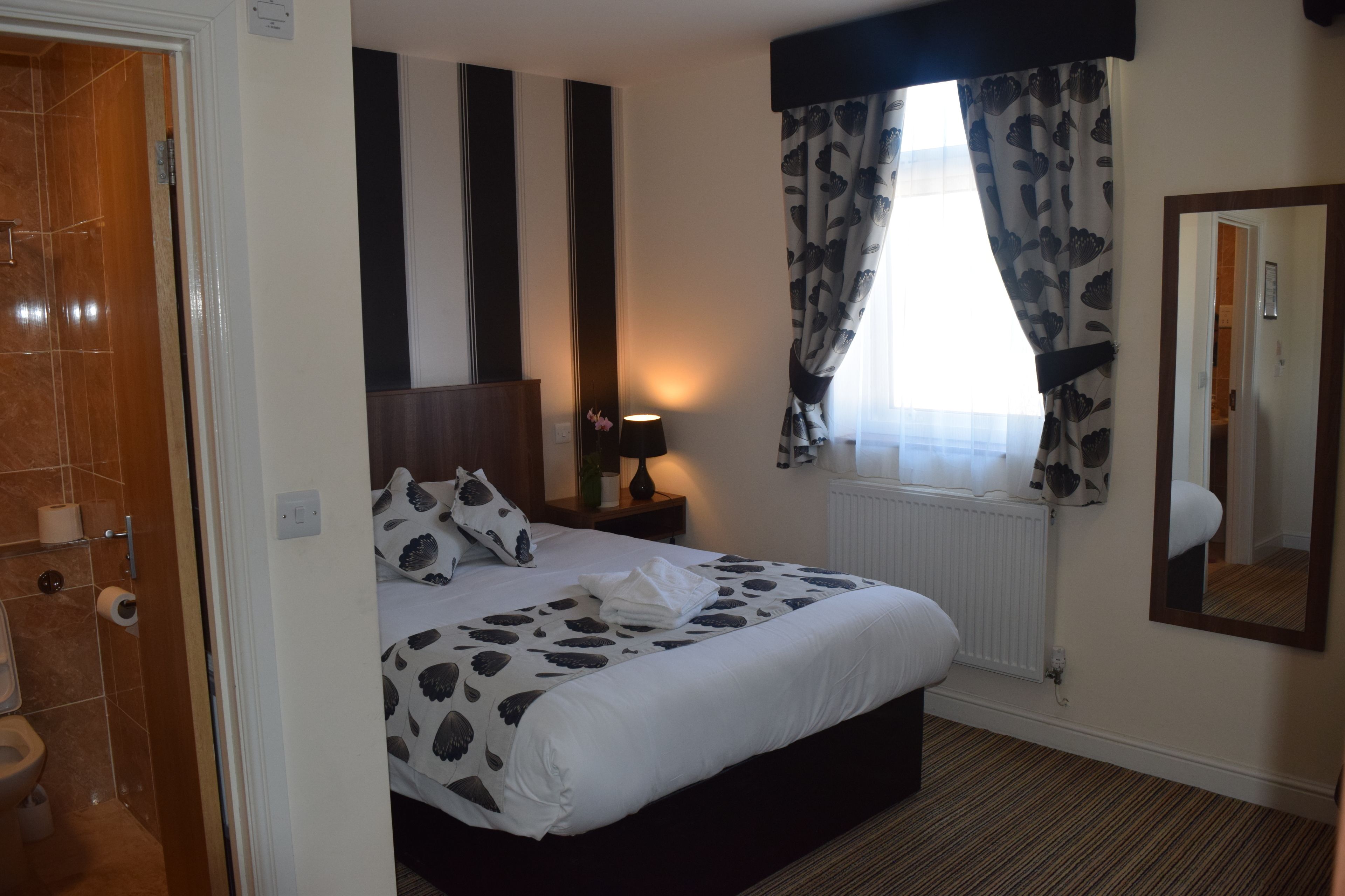 A hotel room with a bed, a nightstand, a lamp, a window with curtains, a radiator, and a mirror on the wall.