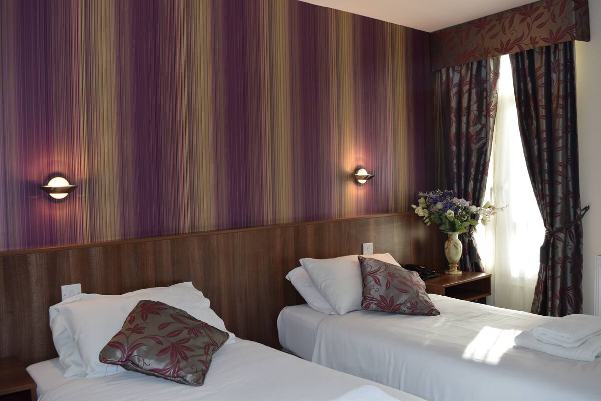 A hotel room with two beds, a wooden headboard, a nightstand with a vase of flowers, and a window with curtains.