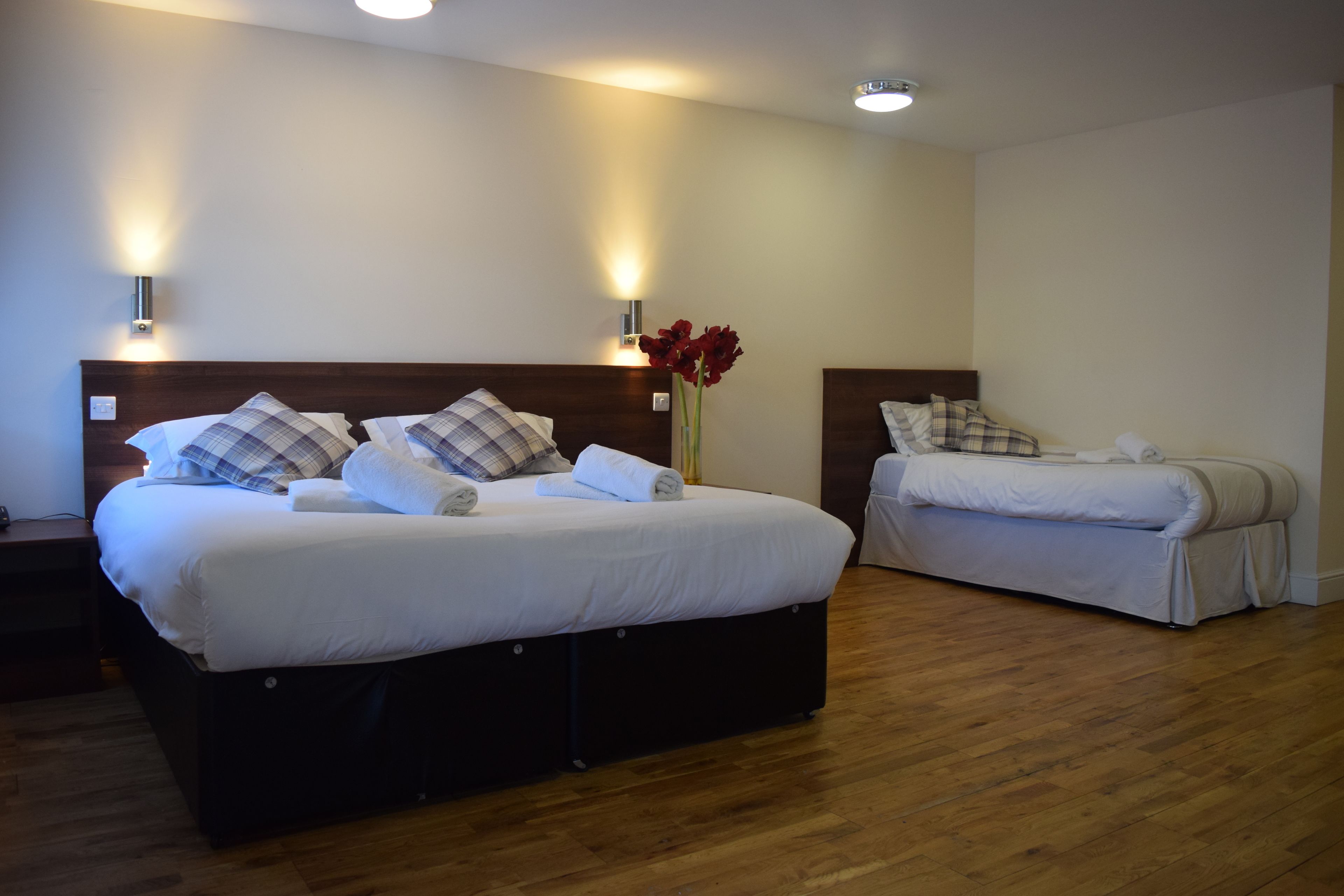 A hotel room with two beds, each with white bedding and plaid pillows. There is a vase with red flowers on the nightstand between the beds.
