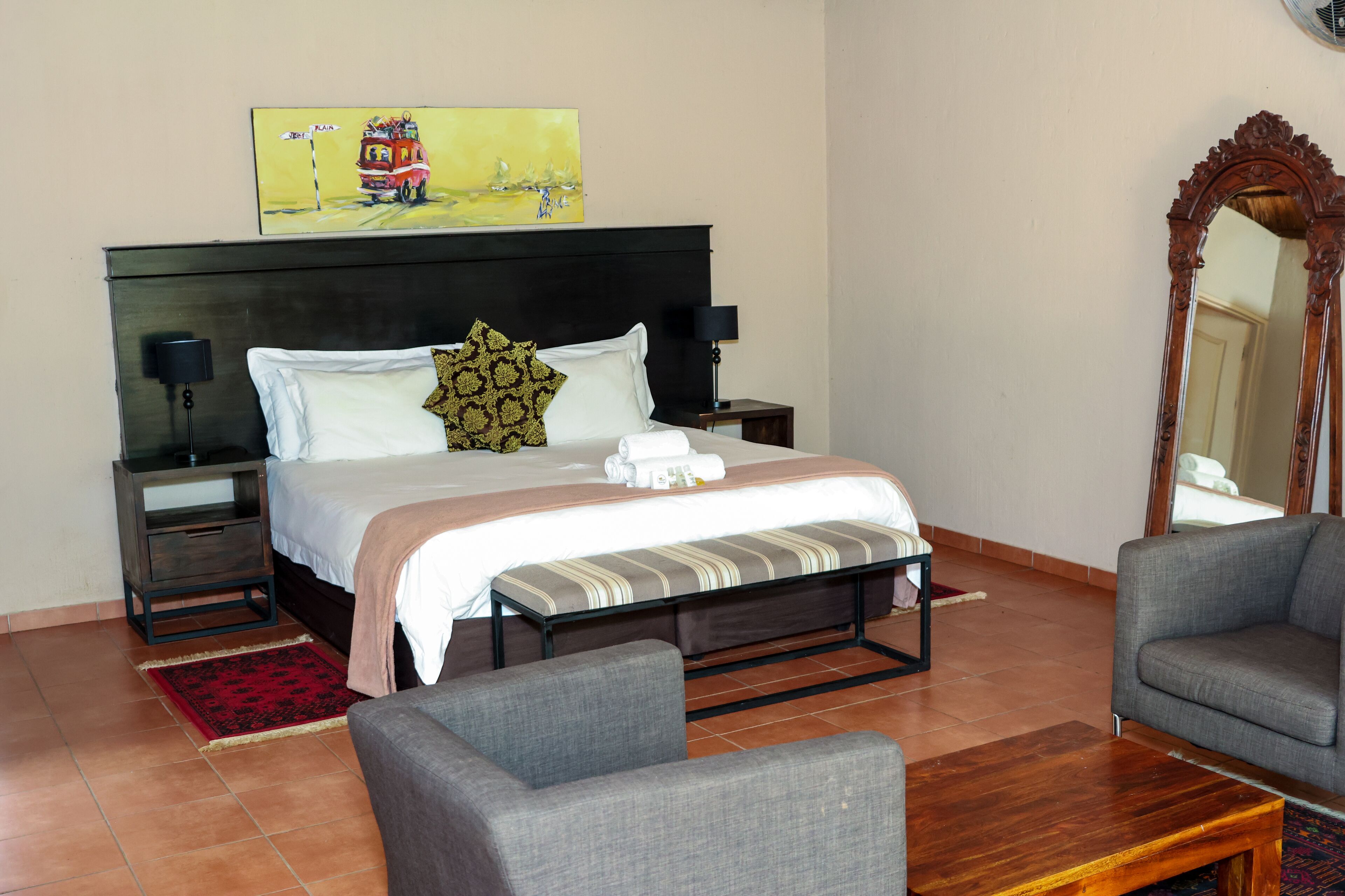 Une chambre d’hôtel avec un grand lit, deux tables de chevet, une petite table, une chaise, une table basse en bois, un miroir et un tableau au mur.