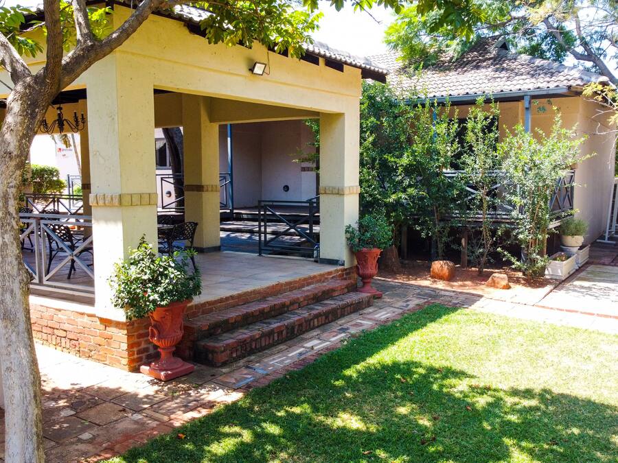 A covered outdoor seating area with a brick pathway leading to a door, surrounded by greenery and trees.
