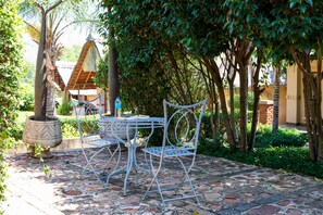 An outdoor patio with a metal table and chairs, surrounded by lush greenery and a stone pathway.