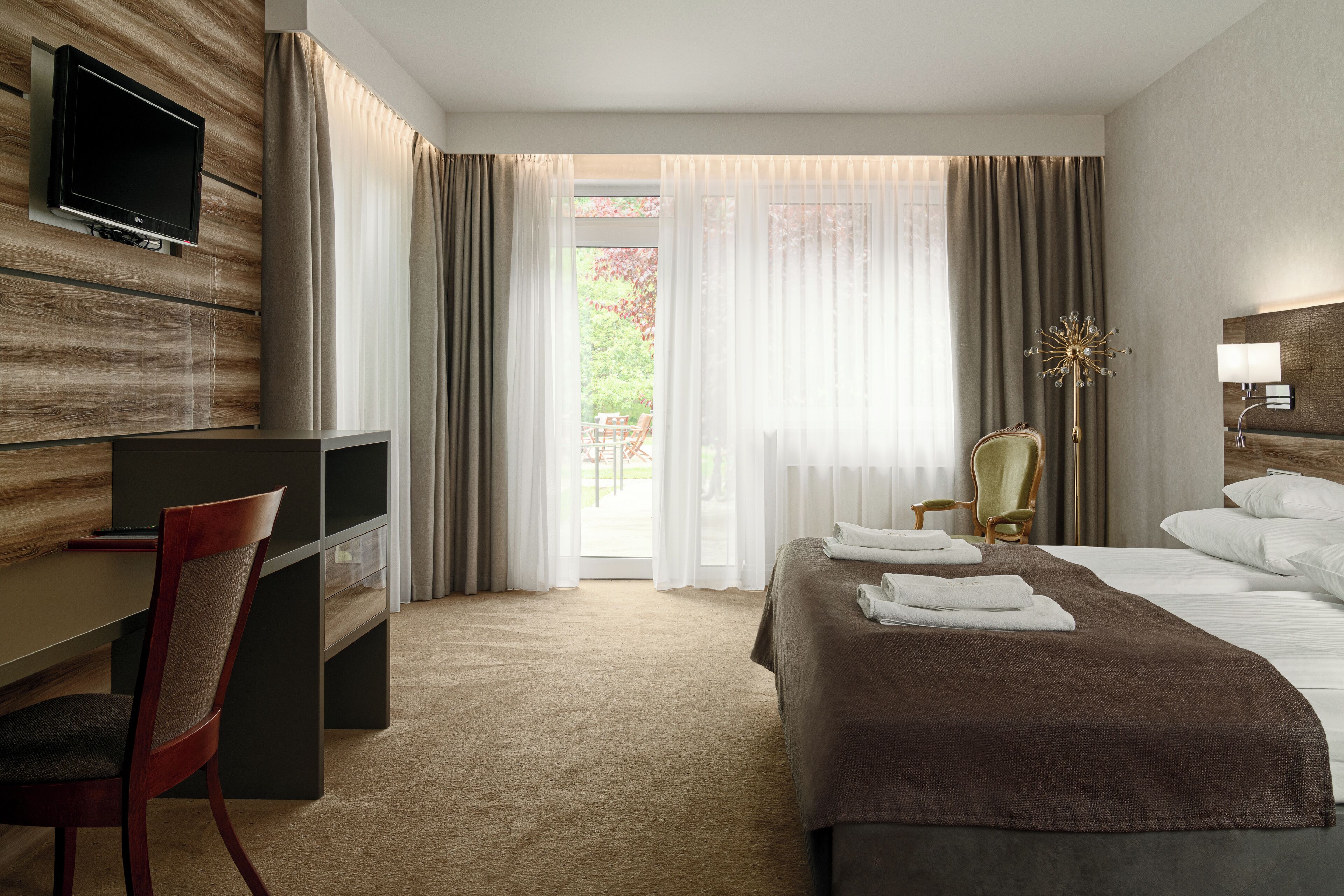 A hotel room with a bed, a desk, a chair, a TV mounted on the wall, and a view of the outdoors.