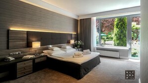 A modern bedroom with a large bed, bedside tables, a lamp, and a view of greenery through large windows. - Villa Eva (Gdansk)