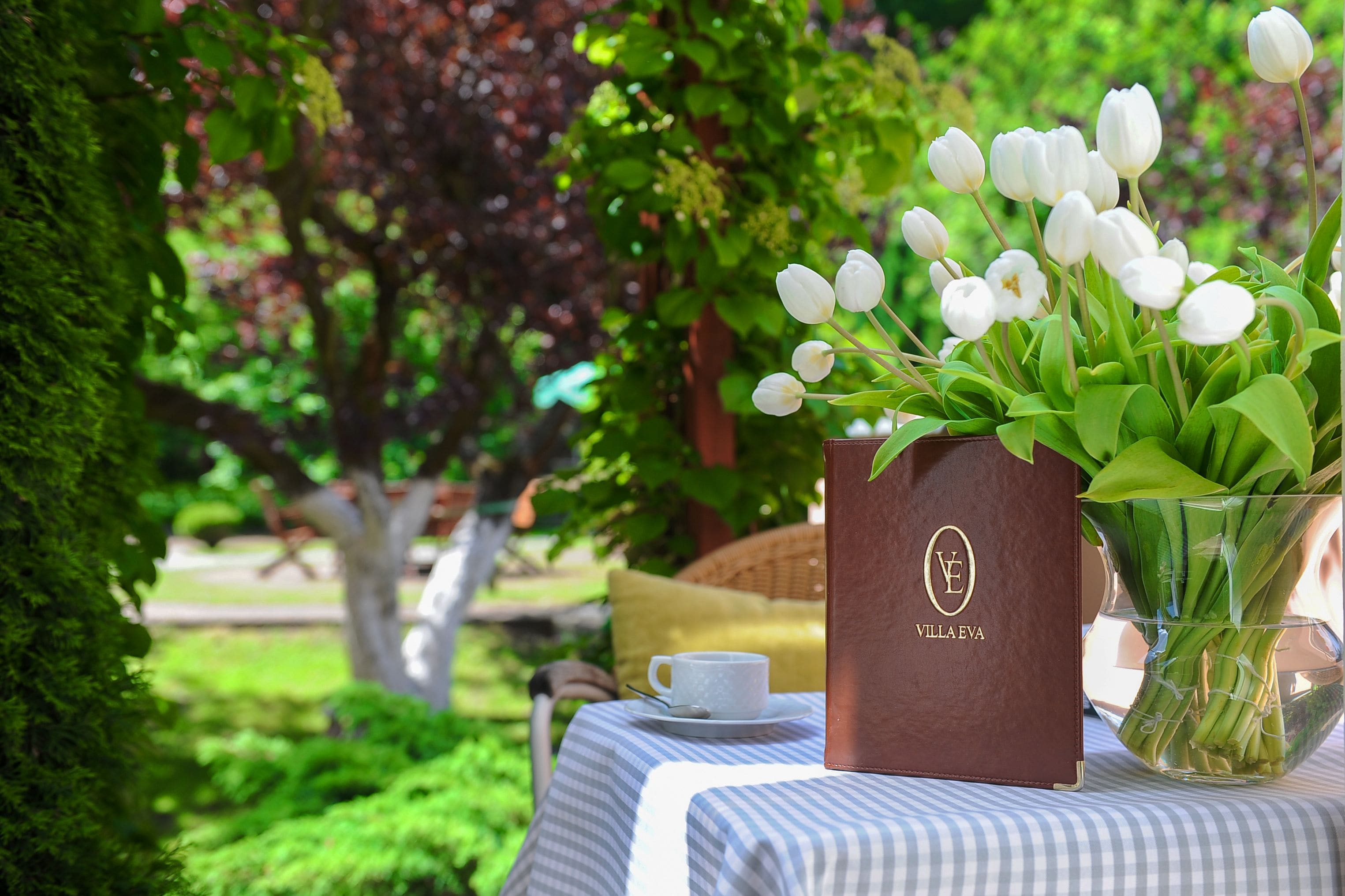 A table with a clear vase of white tulips, a brown leather-bound book, and a white cup on a checkered tablecloth.