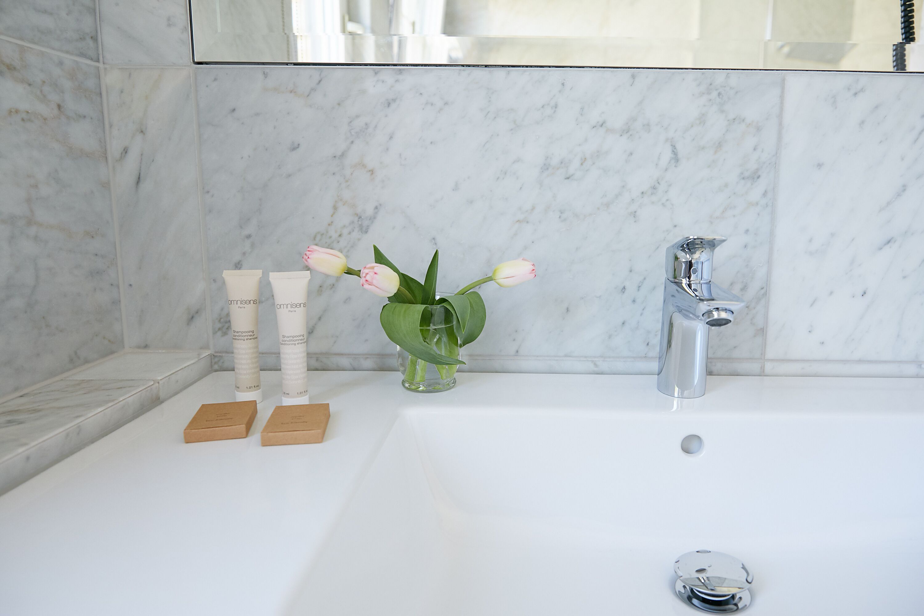 Un baño con pared de mármol, una bañera blanca, un florero de vidrio con flores y dos pequeños cofres de madera.