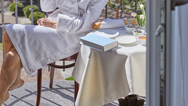 A person seated at an outdoor table with a book, a cup, and a plate, with greenery in the background. - Villa Eva (Gdansk)