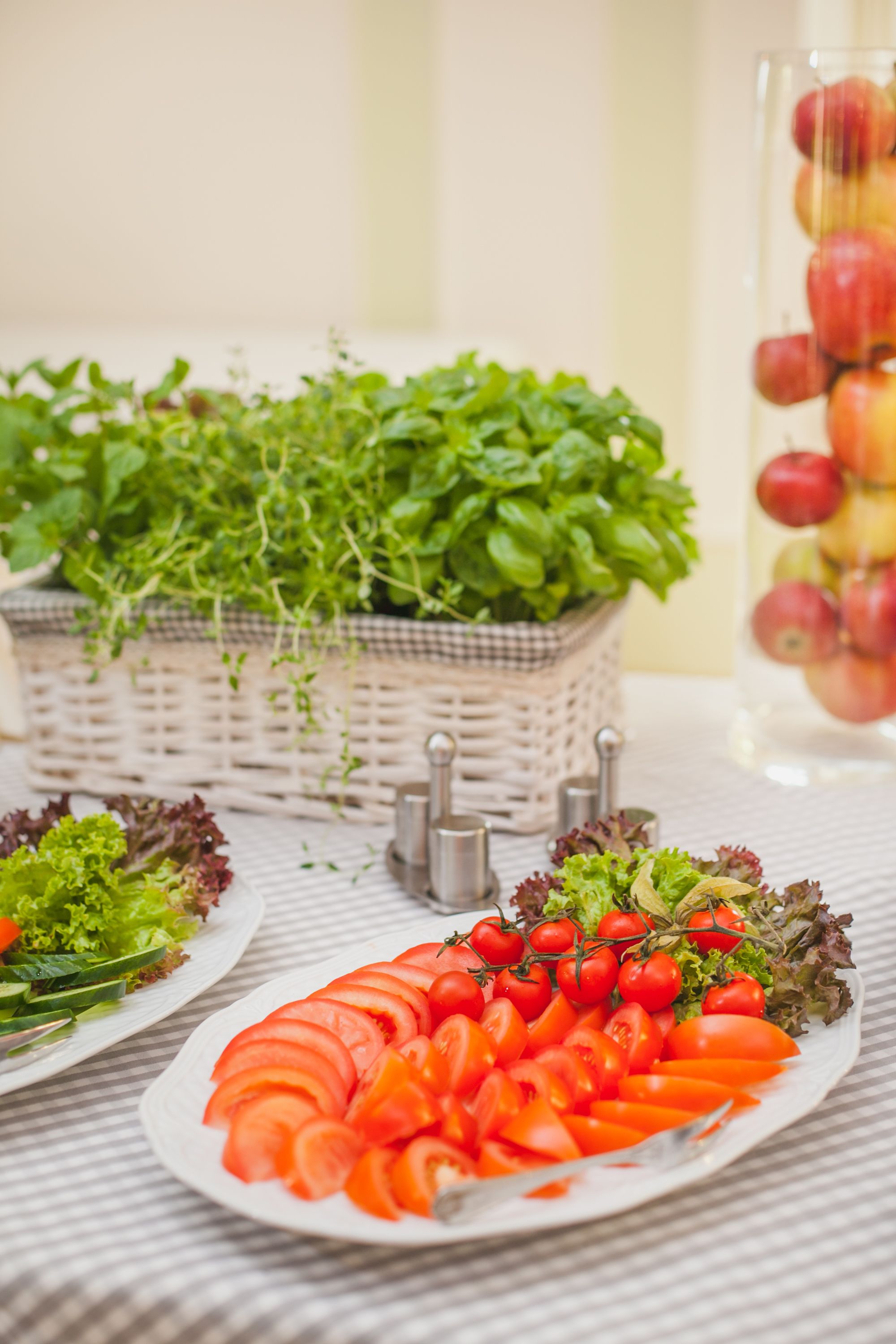 Un tavolo con piatti di pomodori a fette e insalata, un cesto di erbe aromatiche e un vaso di vetro con delle mele.