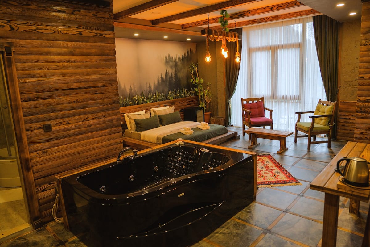 A log cabin-style room with a large black bathtub, a sofa with pillows, a wooden table with a kettle, and a chair with a cushion.