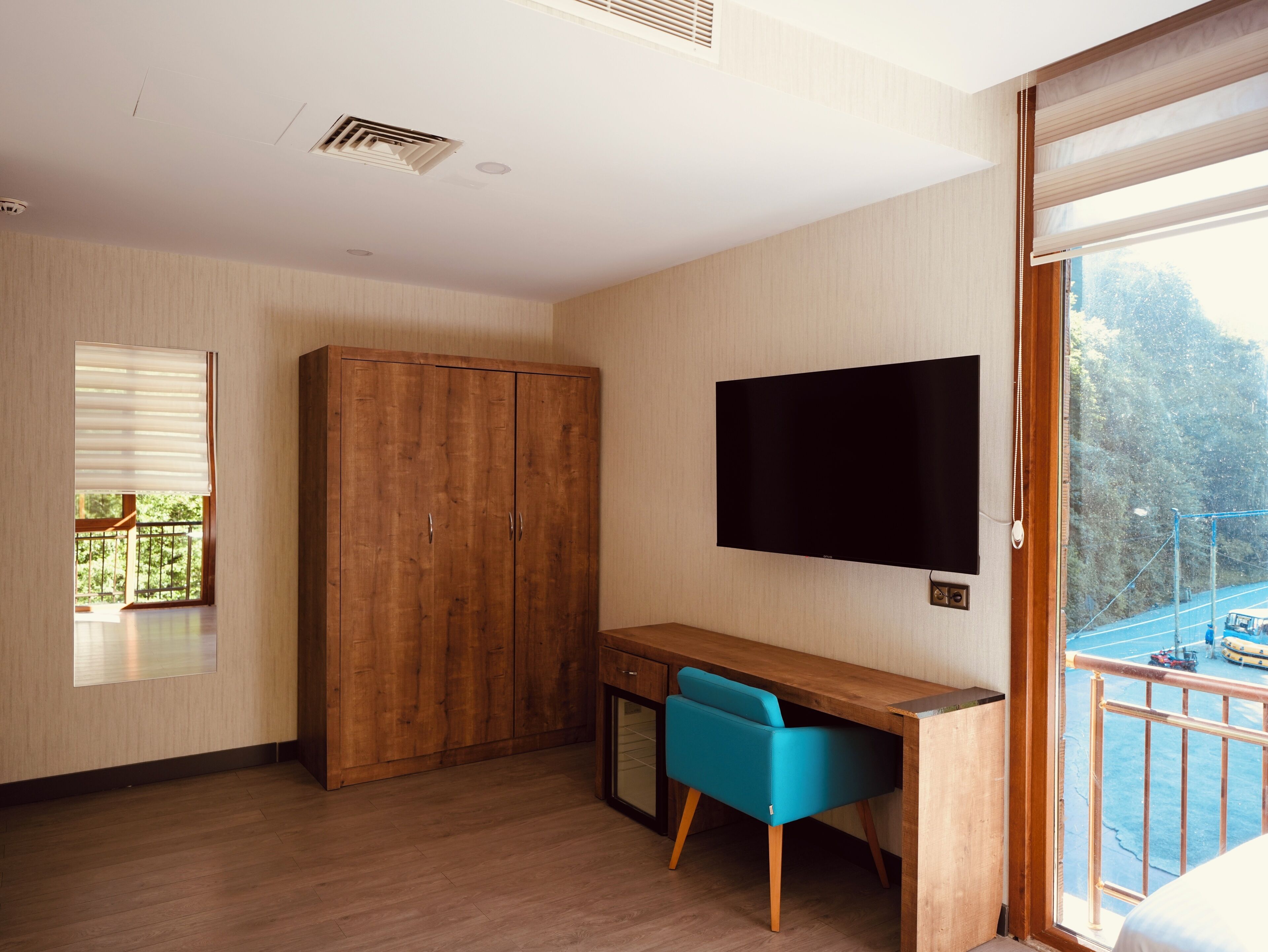 A room with a wooden wardrobe, a desk with a chair, a mounted TV, and a balcony with a view of trees and a road.