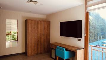 A room with a wooden wardrobe, a desk with a chair, a mounted TV, and a balcony with a view of trees and a road.