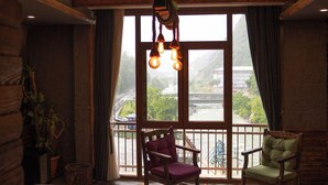 A cozy room with a bed, two chairs, a small table, and a view of a river through large windows. - Ayder Hanedan Hotel (Ardesen)