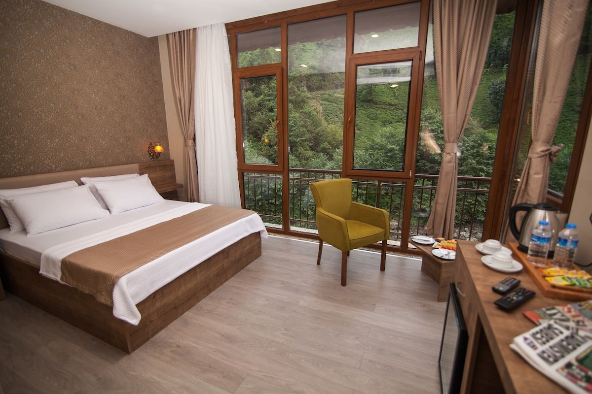 A hotel room with a bed, a yellow armchair, a dining table with breakfast items, and large windows with a view of greenery.
