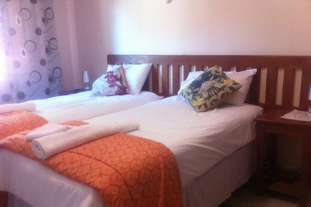 A hotel room with two beds, each with a wooden headboard, white linens, and orange patterned bedspreads. There is a nightstand with a lamp and a floral pillow on one bed.