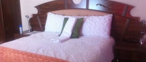 A neatly made bed with a patterned blanket, a wooden headboard, and a bedside table with a lamp and a bottle.