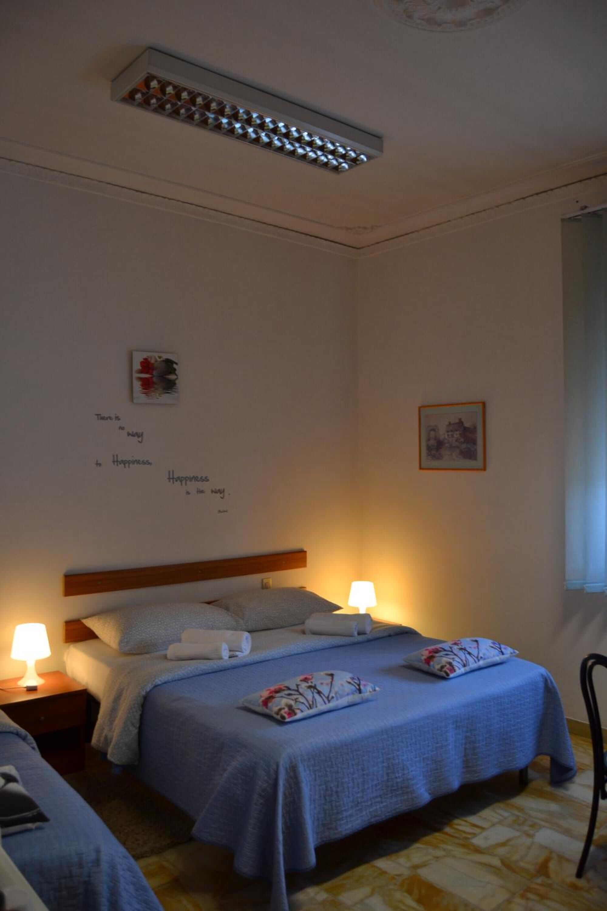 A bedroom with a bed, two bedside tables with lamps, a ceiling light fixture, and a wall with a framed picture and text.