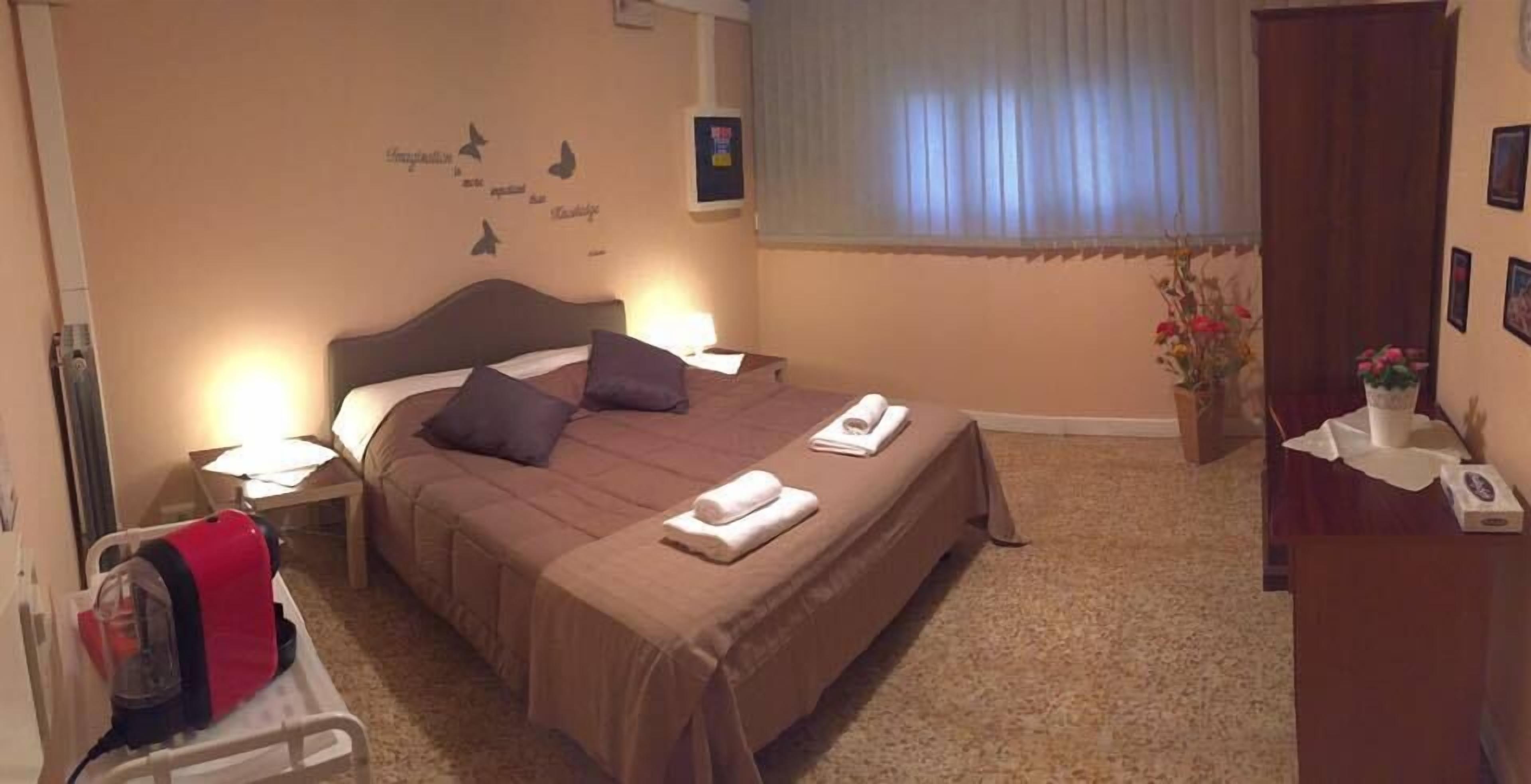 Una cama bien hecha con almohadas, una mesita de noche con una lámpara, una pequeña mesita de noche con un libro, una pared con un adhesivo de mariposa y una ventana con persianas.