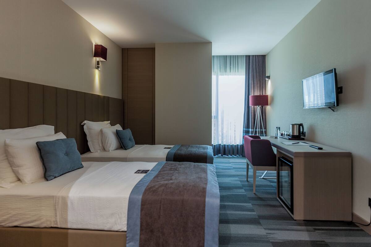 Ein Hotelzimmer mit zwei Betten, einem Schreibtisch, einem Stuhl, einem Fernseher und einem Fenster mit Vorhängen.