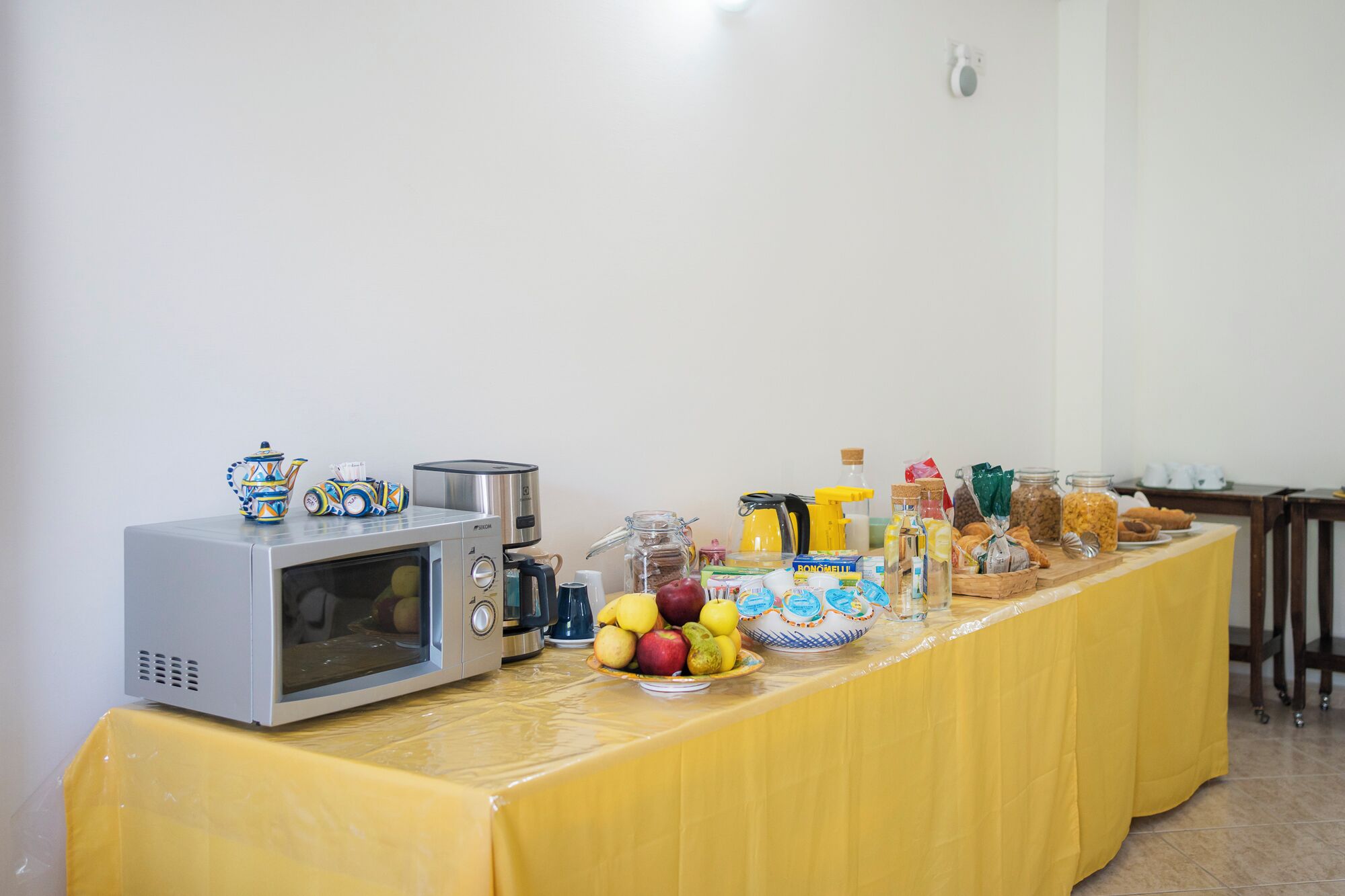 Una mesa con mantel amarillo presenta una barra de desayuno con microondas, cafetera y diversos alimentos.