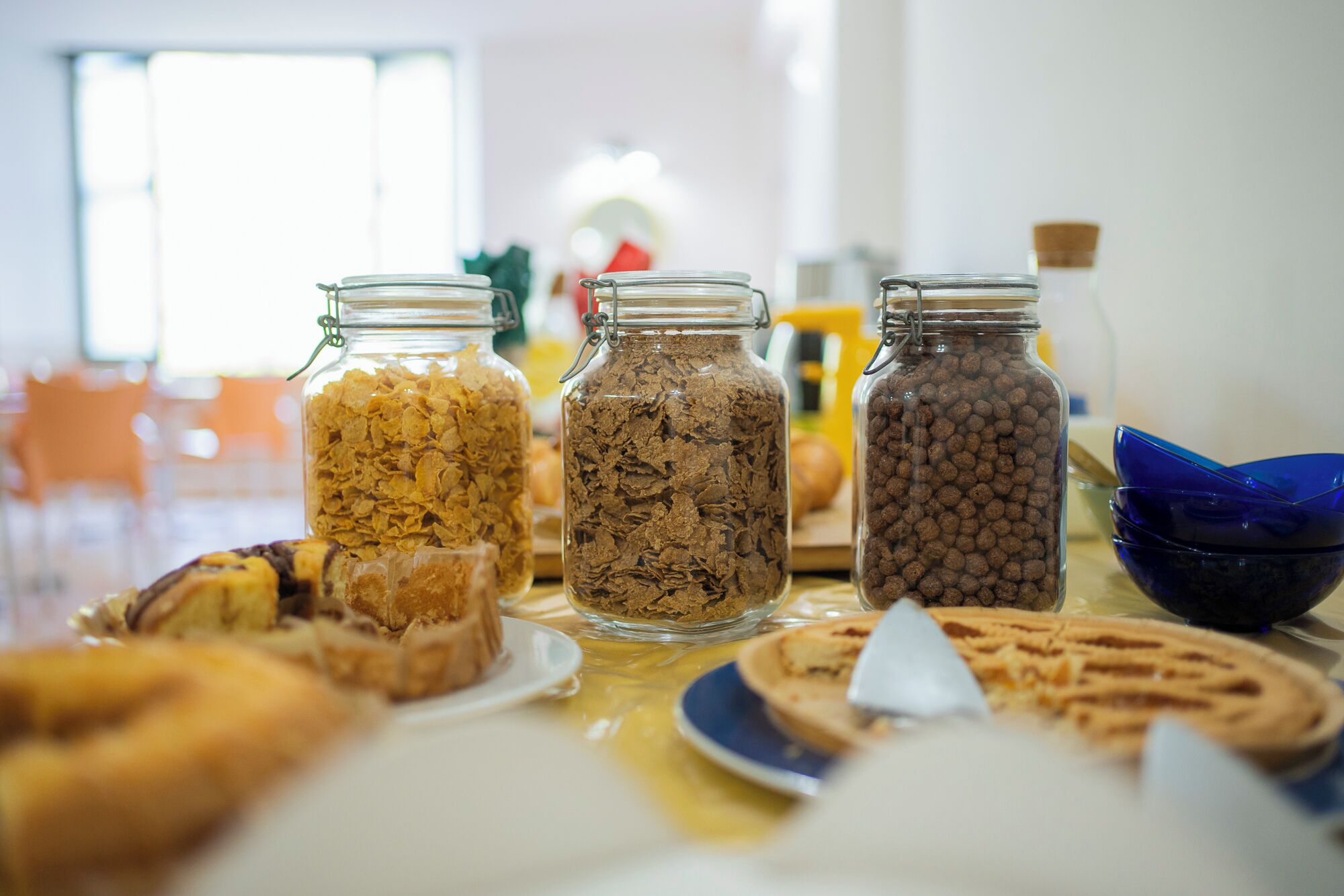 Una mesa con tres frascos de vidrio llenos de diferentes tipos de cereal, un gofre y un pastel sobre un mantel.