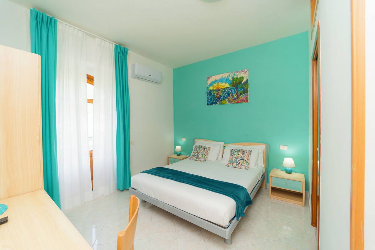 A bedroom with a bed, two bedside tables, a chair, a window with curtains, and a painting on the wall.