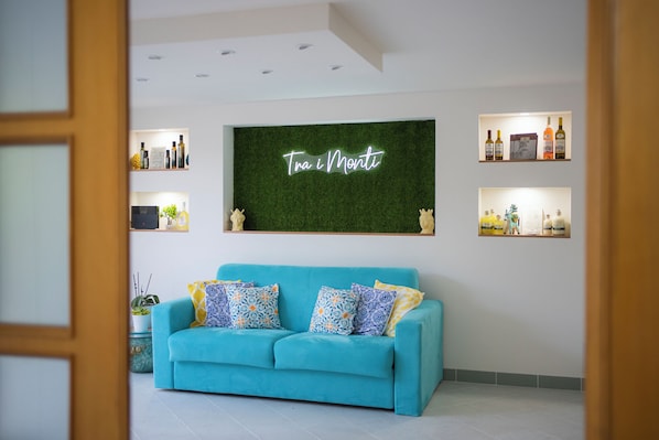 A modern living room with a teal sofa, decorative pillows, a green wall with a neon sign, and shelves with bottles and decorative items.
