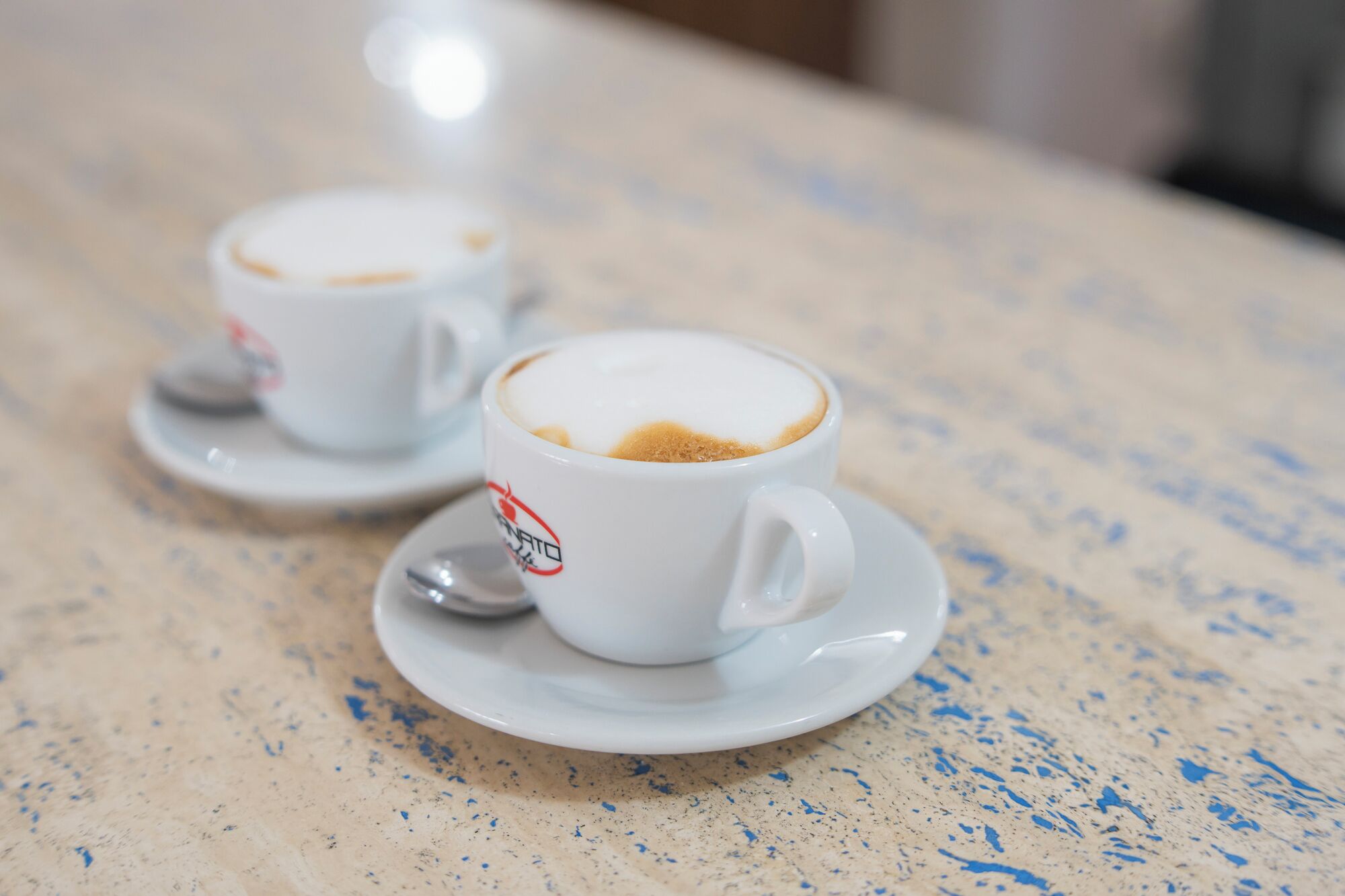 Zwei weiße Kaffeetassen mit einem Logo auf dem Untersetzer, auf einer fleckigen Arbeitsplatte platziert.